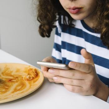 A imagem mostra uma menina  sentada à mesa usando um smartphone logo após comer um prato de macarrão.