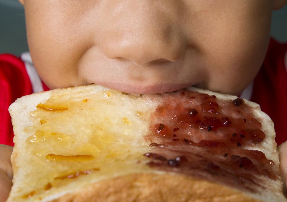 A parte da boca e do nariz do rosto de uma criança aparece na foto. A criança morte um pão com geleia, ilustrando a presença dos carboidratos na alimentação infantil