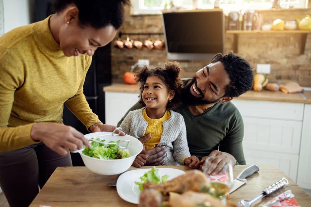 Mãe serve salada no prato para a sua filha, em busca de fortalecer a imunidade. A criança está sentada no colo do pai e todos sorriem.