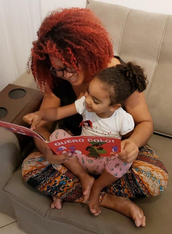 Monique Gonçalves lê livros para a sua filha. A leitura é de um livro chamado "Quero Colo!" e as duas estão sentadas no sofá.