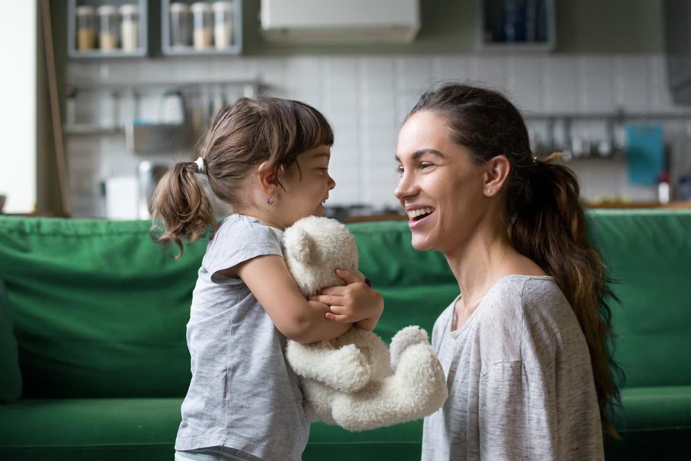 Mãe e filha se olham sorrindo, em frente a um sofá, na sala de casa. A mãe está abaixada no mesmo nível da menina, que abraça um ursinho de pelúcia.