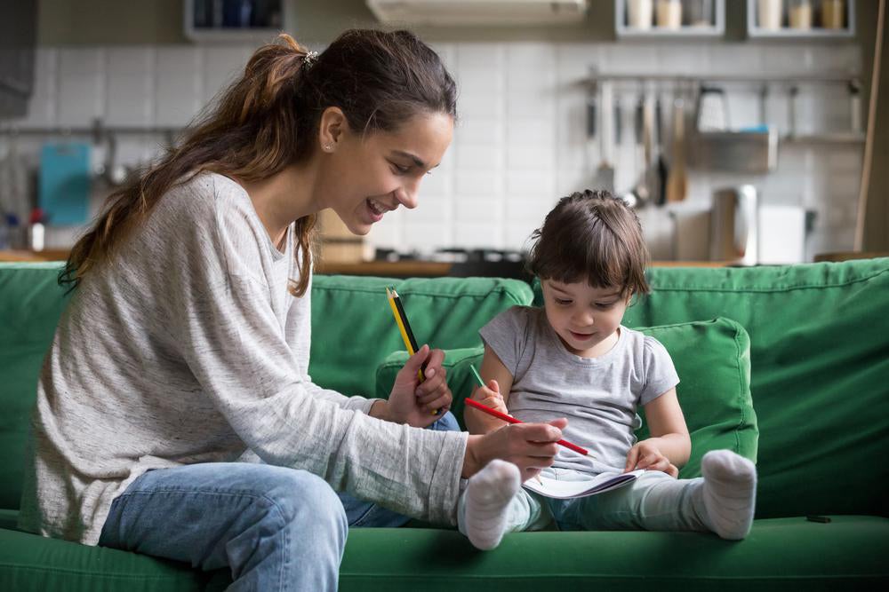 Uma babá está sentada no sofá ao lado de uma criança. Ela ajuda a garotinha a desenhar e pintar. 