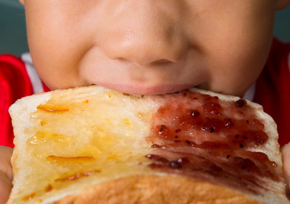 A parte da boca e do nariz do rosto de uma criança aparece na foto. A criança morte um pão com geleia, ilustrando a presença dos carboidratos na alimentação infantil