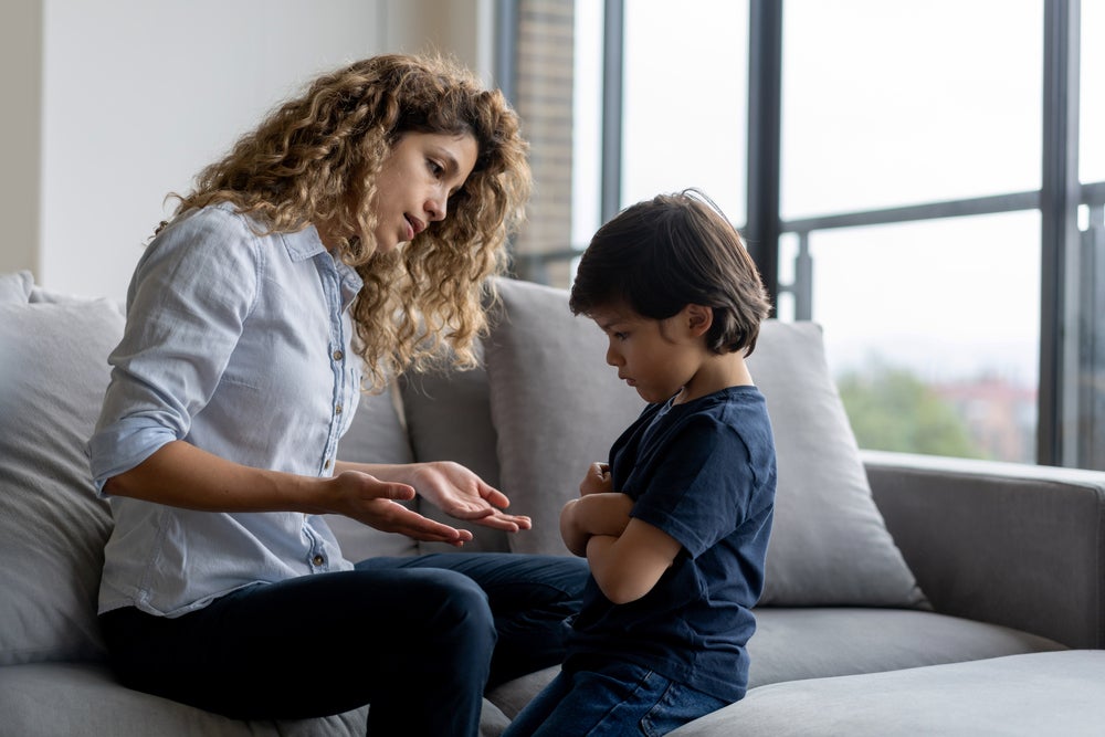 Uma mãe está sentada em um sofá e conversa com seu filho, representando como dizer não. O filho está em sua frente, com braços fechados e rosto de insatisfação. 