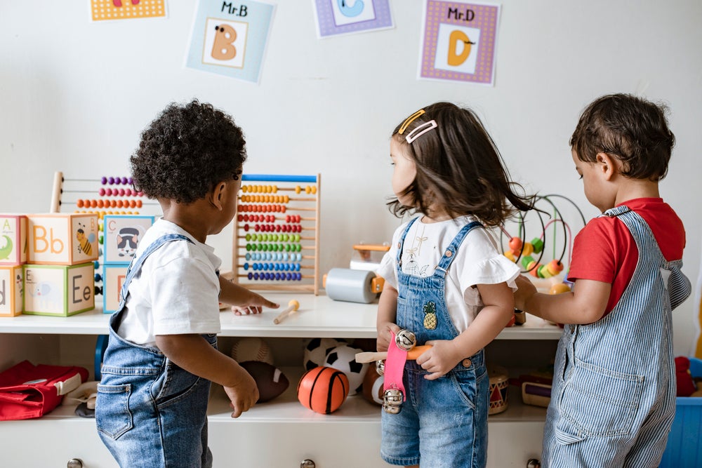 Três crianças brincam com brinquedos diversos dentro de uma sala de creche.