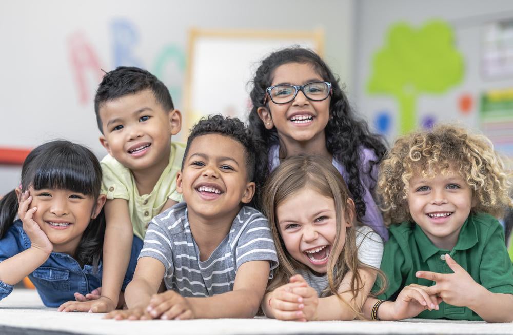 Seis crianças, garotos e garotas, posam para a foto sorrindo em um cenário de sala de aula, ilustrando a diversidade de gênero. 