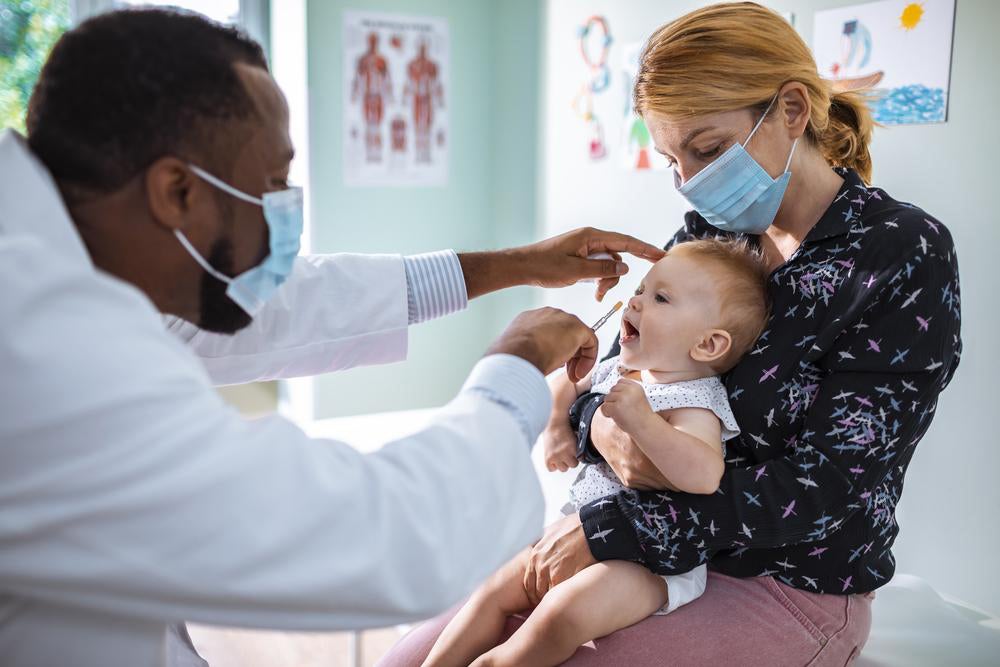 Em um consultório, um médico pediatra examina um bebê que está no colo de sua mãe. 