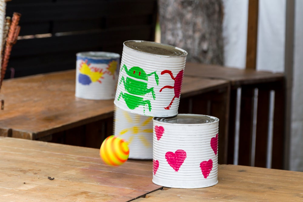 Artesanato feito em lata de leite Ninho. São quatro latas pintadas com tinta branca e com desenhos coloridos, todas apoiadas em uma mesa de madeira. 