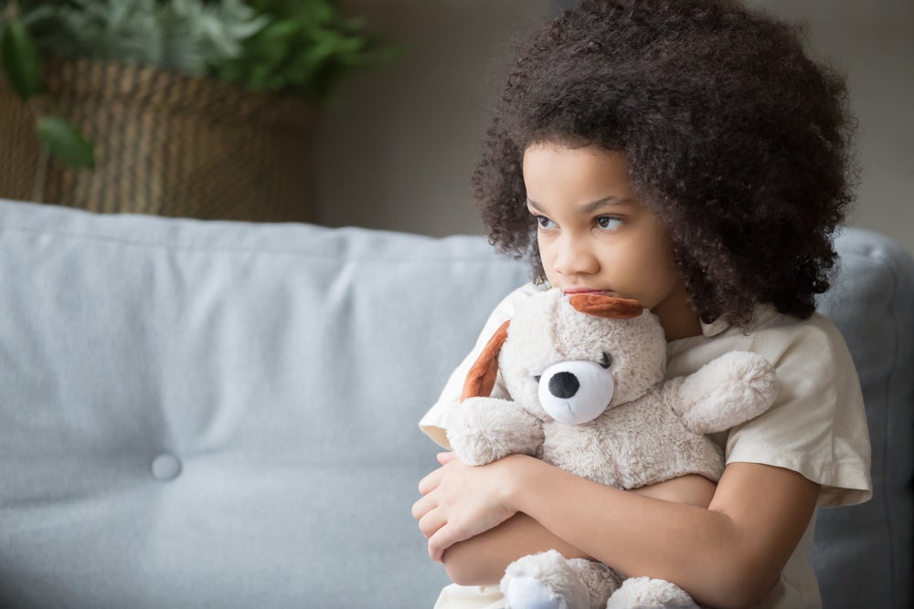 Uma criança abraça um urso de pelúcia com o rosto triste, ilustrando a atenção para a saúde mental infantil. 