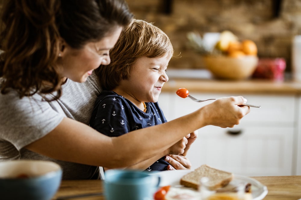 A seletividade alimentar é ilustrada pela foto de uma mãe tentando dar um tomate com um garfo para a sua criança, que está sentada em seu colo. A criança está de olhos e boca fechada, se negando a comer. 