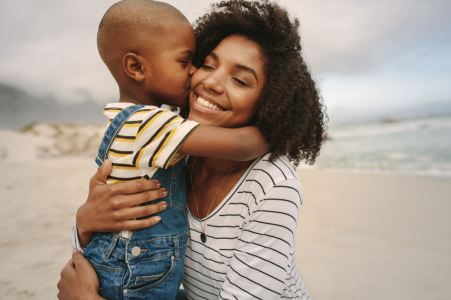 Uma mãe está ajoelhada na areia e recebe um abraço e um beijo de seu filho. Ela está com os braços ao redor dele, sorrindo de olhos fechados, e representa um dos diferentes tipos de mãe na relação com os filhos. 