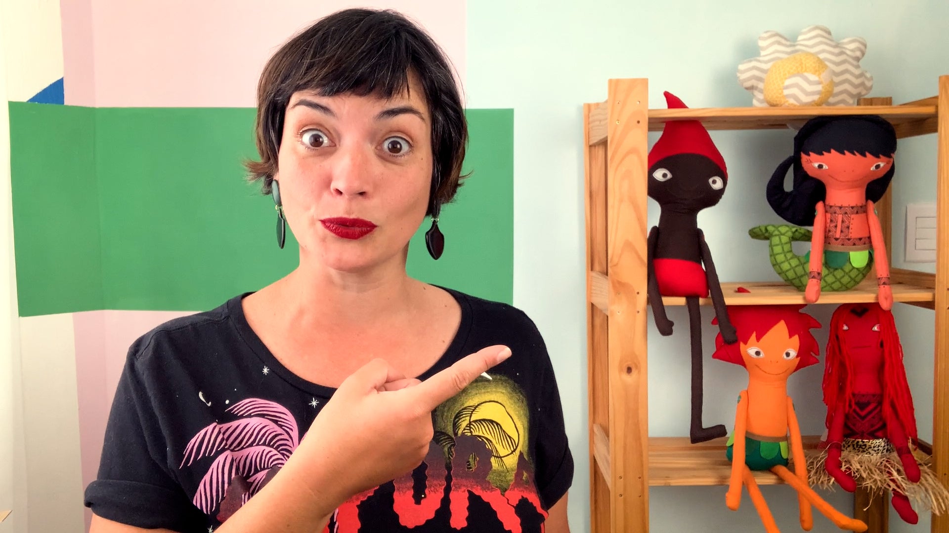 Foto de Flávia Scherner, do canal Fafá Conta Histórias. Ela está apontando para um estante com bonecos de pano que representam personagens do folclore brasileiro.