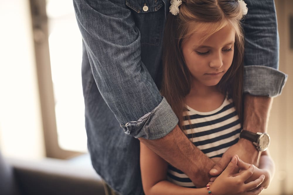 Na imagem, há um homem e uma criança. O homem usa casaco jeans e relógio. Ele está abraçado com uma criança, por trás, e os dois estão de mãos dadas, uma forma de acalmar a ansiedade em crianças. A menina usa blusa listrada e flores brancas no cabelo