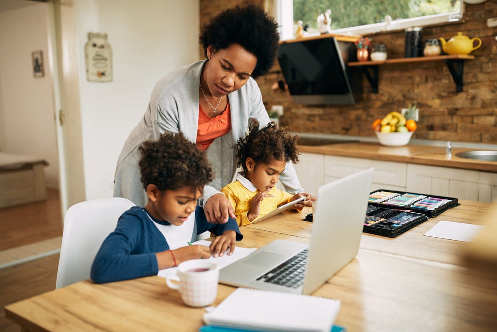 Em um ambiente de casa, uma mãe auxilia seu filho e sua filha em atividades de aulas remotas.