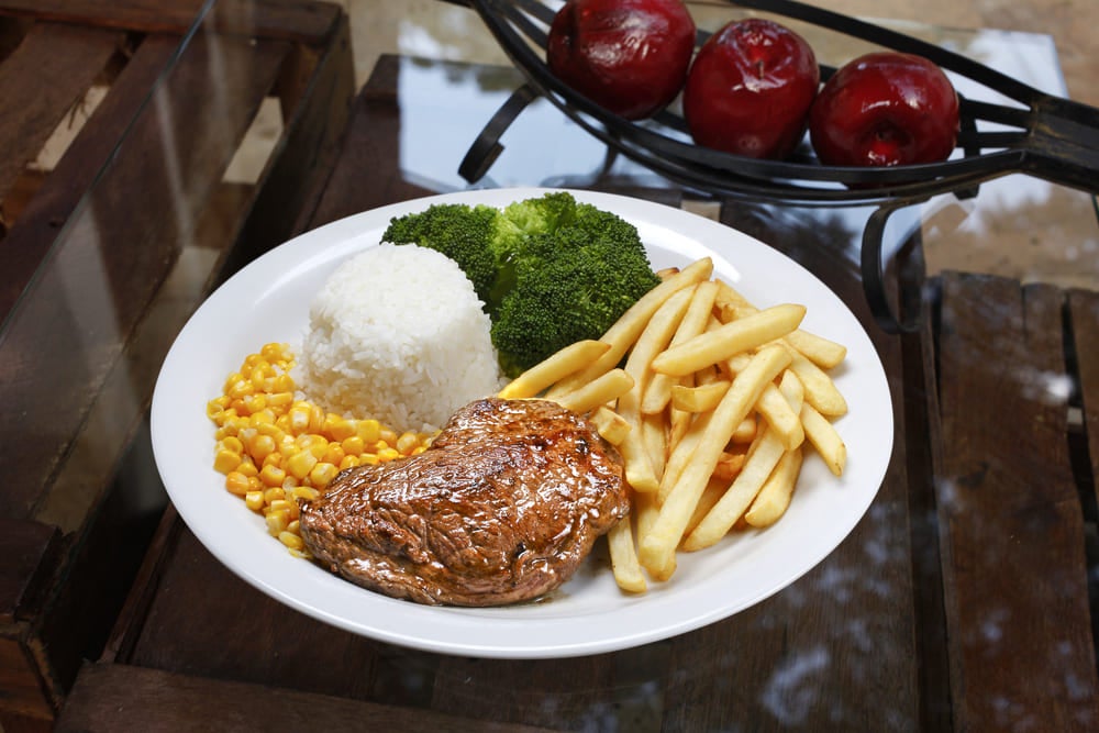 Sobre uma mesa estão um arranjo com três maçãs e um prato com arroz, milho, carne, brócolis e batatas fritas. Representam as comidas típicas do Brasil.