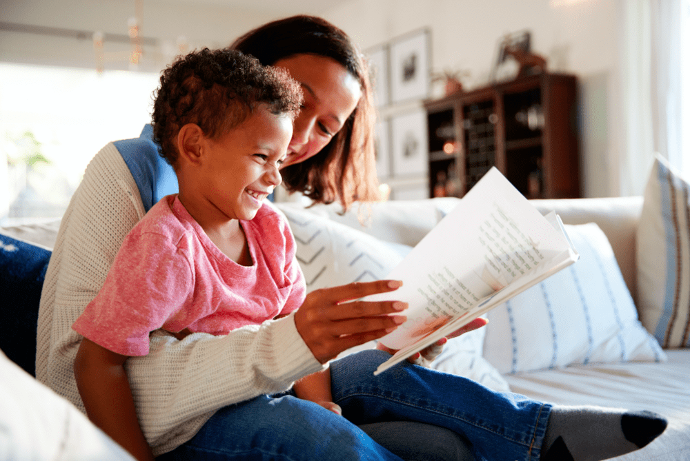 Mãe sorri com o filho sentado em seu colo enquanto os dois leem um livro juntos representando a ideia da mãe suficientemente boa.