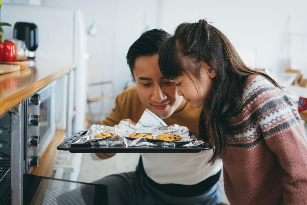 Um pai e sua filha estão em frente a um forno segurando uma bandeija de cookies na cozinha, como exemplo de atividades de dia dos pais. 