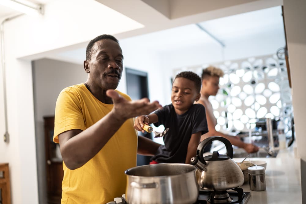 Pai e filho cozinham juntos como uma homenagem de dia dos pais. O pai está mexendo uma panela e o filho, ao seu lado, o ajuda. 