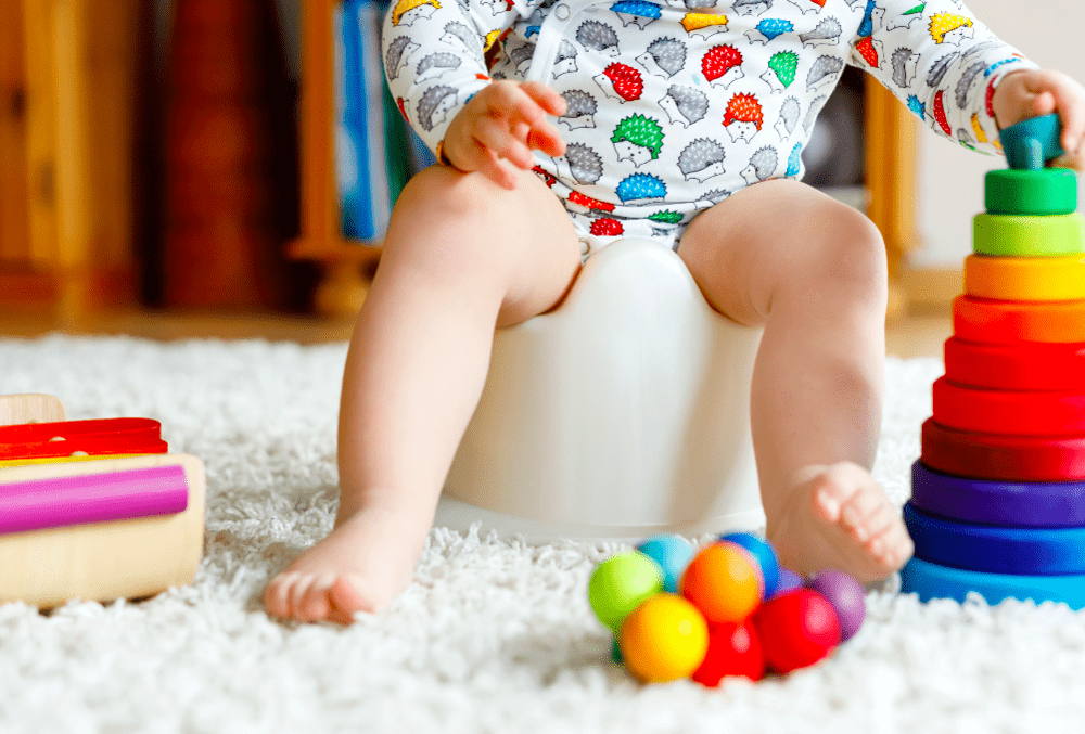 Criança sentada em um penico para fazer cocô, cercada de brinquedos coloridos.