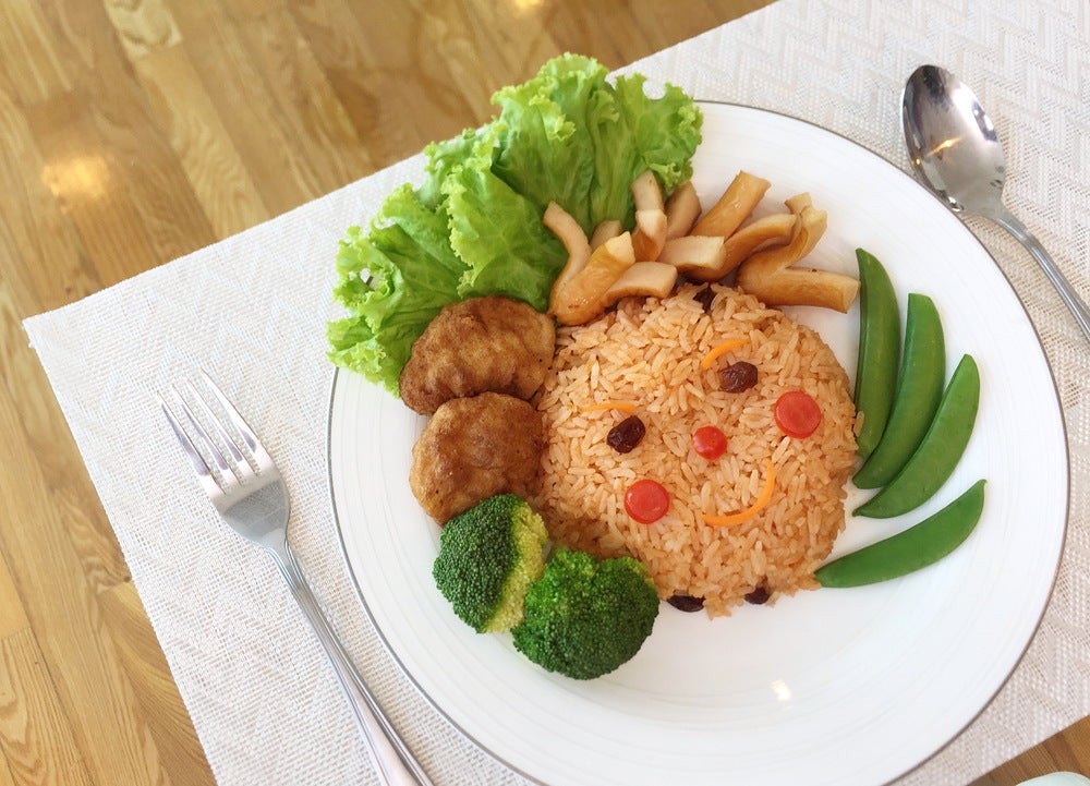 Um prato com arroz, alface, brócolis e outros alimentos monta uma carinha. Ele está sobre uma mesa e, ao lado esquerdo há um garfo, enquanto ao direito uma colher