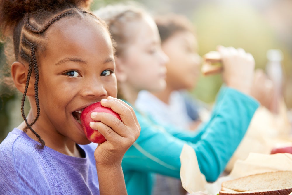 Na imagem, há uma menina mordendo uma maçã vermelha e duas crianças ao seu lado. Todas estão sentadas e ilustram a alimentação saudável infantil.