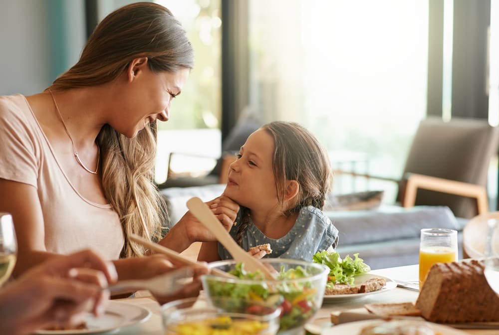 Na imagem, há uma mulher e uma menina sentadas em frente à mesa de almoço, em uma sala de estar. A mulher está olhando carinhosamente para a menina, que, por sua vez, está sorrindo para ela. Nos pratos, há alface com legumes, fatias de pão integral e um suco de laranja