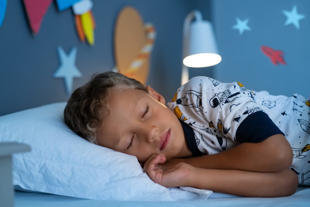 Um menino, vestido de pijama estampado, está deitado na cama, de olhos fechados. O abajur atrás dele está aceso, e nas paredes do quarto há decorações de planetas. 