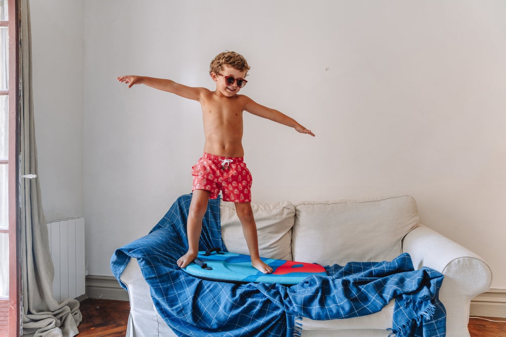 Um menino está brincando de surf em cima do sofá O menino veste roupas de banho e óculos escuros. Ele está em cima de uma prancha de surf, que por si só está em cima de uma manta azul que cobre o sofá, simulando uma onda. 