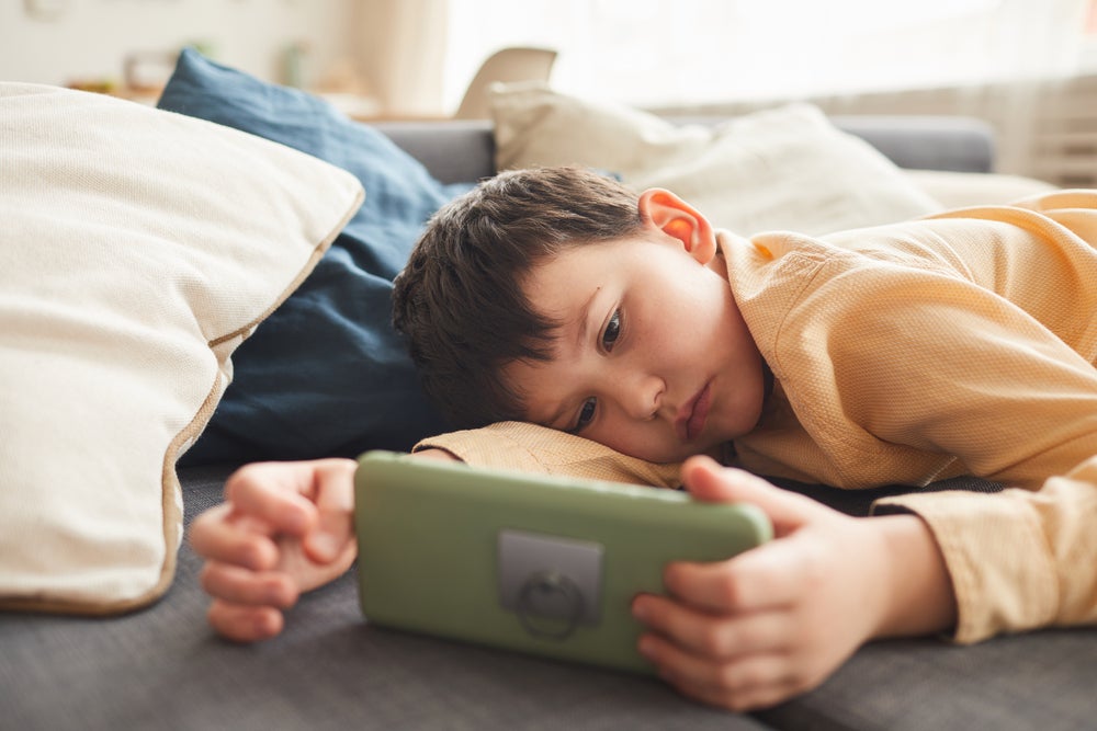 Uma criança está deitada com rosto sério no sofá, olhando para o celular que está em suas mãos, representando o sedentarismo infantil.