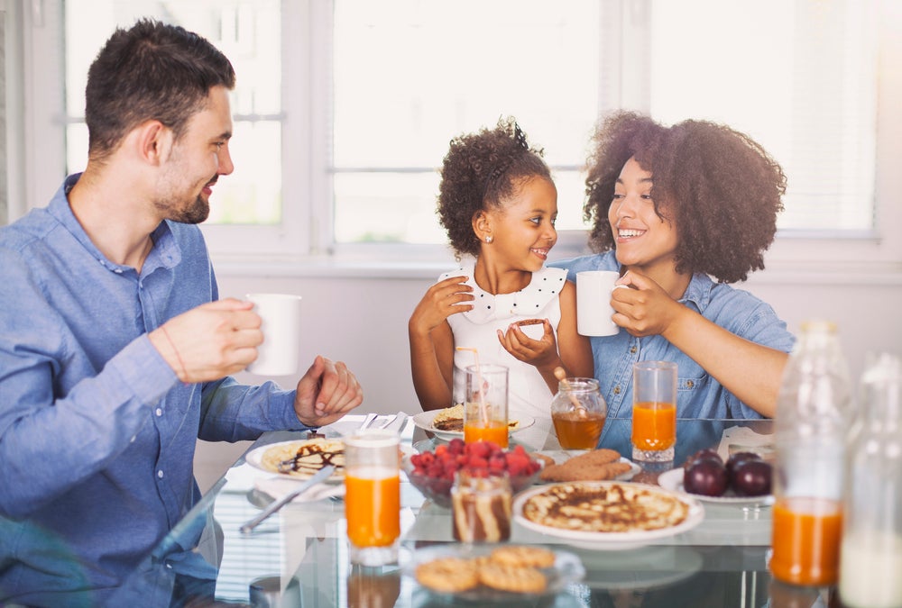 Foto de uma família composta por pai, mãe e criança. Eles estão sorrindo, sentados em frente a uma mesa, onde há um café da manhã nutritivo