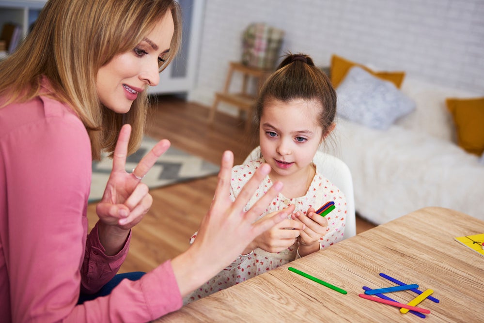 Uma mulher mostra números com os dedos, prática comum no ensino de matemática para crianças, para uma menina que a observa. 