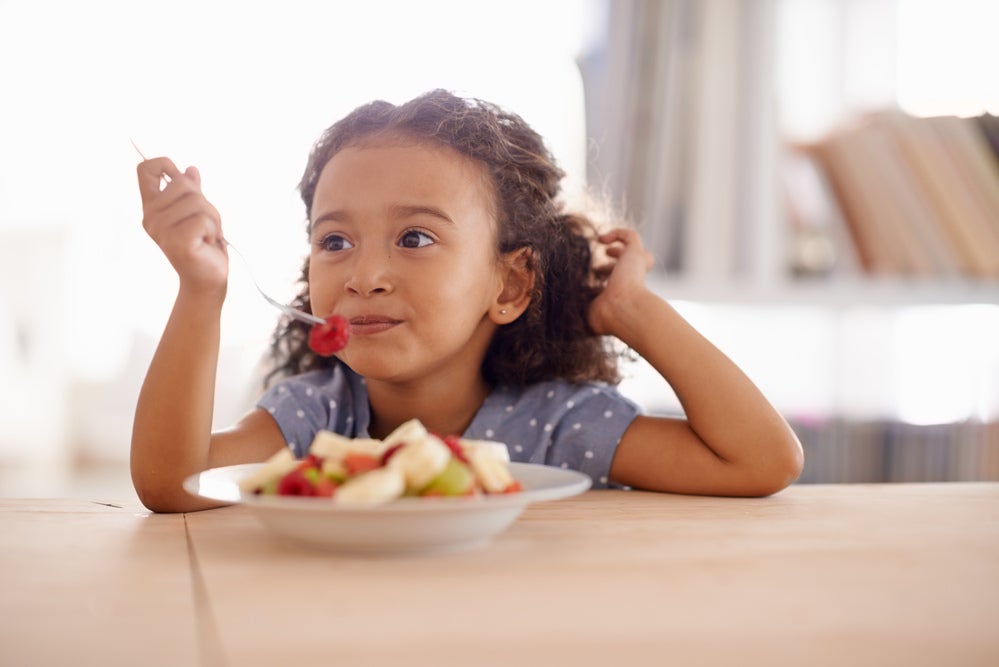 Criança sentada à mesa comendo um prato de frutas, representando uma refeição saudável