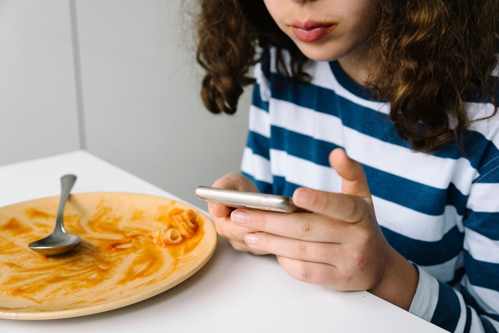 A imagem mostra uma menina  sentada à mesa usando um smartphone logo após comer um prato de macarrão.