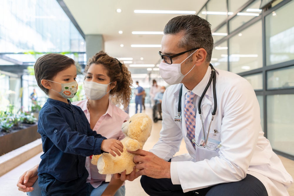 Neuropediatra e enfermeira se aproximam de uma criança, que segura um urso de pelúcia. 