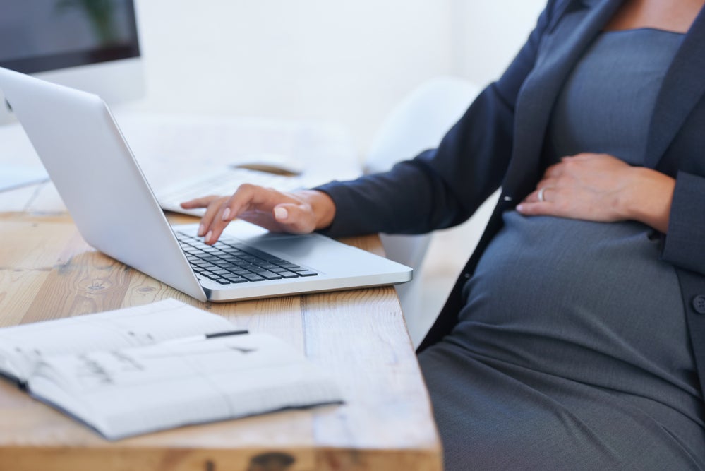Uma mulher grávida digita no computador em um escritório, com uma das mãos sobre a barriga. 