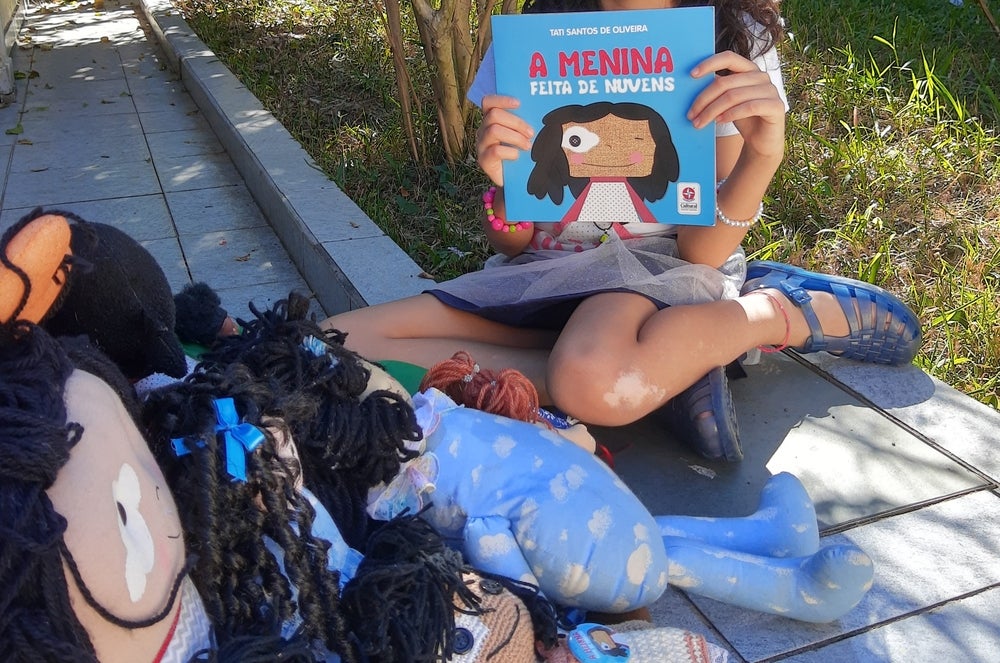 Uma menina com vitiligo sentada no chão, segurando o livro “A menina feita de nuvens”. Ao redor, há várias bonecas parecidas com ela.