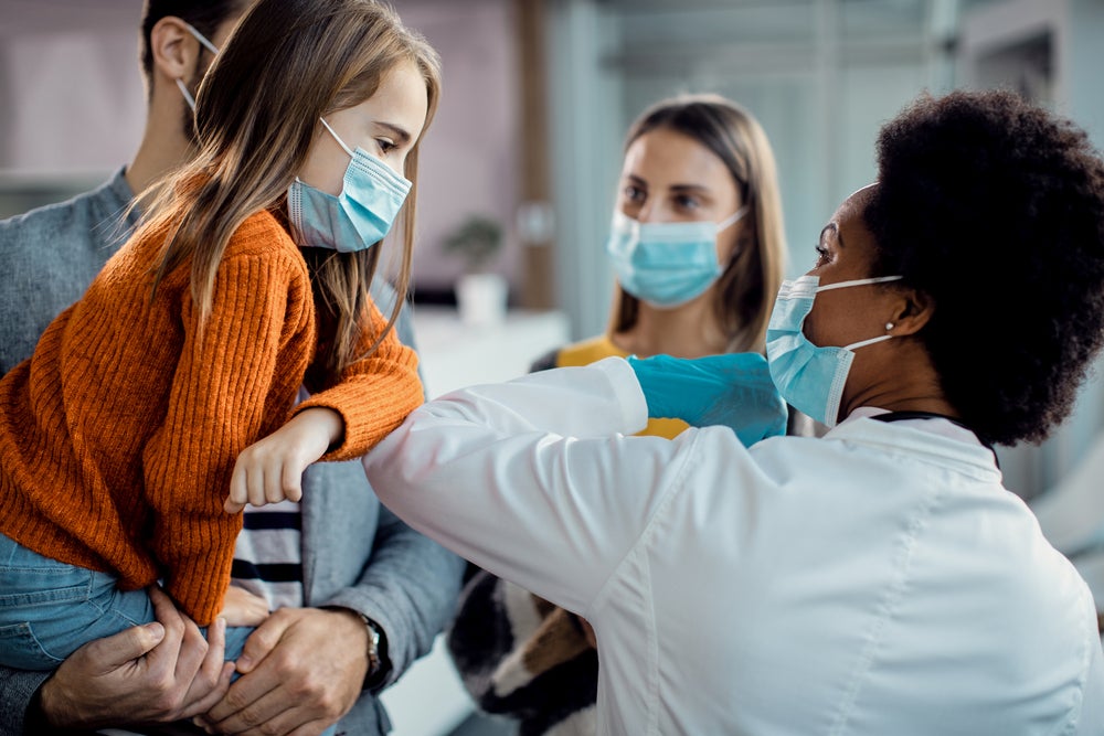 A foto mostra uma criança no colo do pai, junto com a sua mãe em um ambiente hospitalar com uma médica 