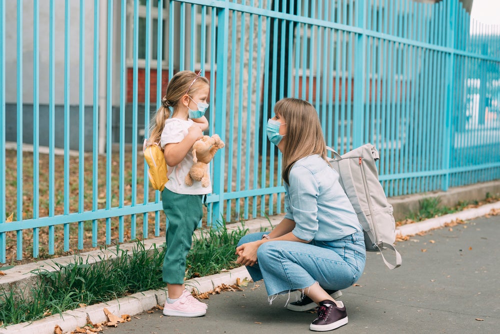 Uma mãe está agachada em frente a sua filha, perto de um portão. A menina está de mochila, olhando para a mãe e segurando um urso de pelúcia.