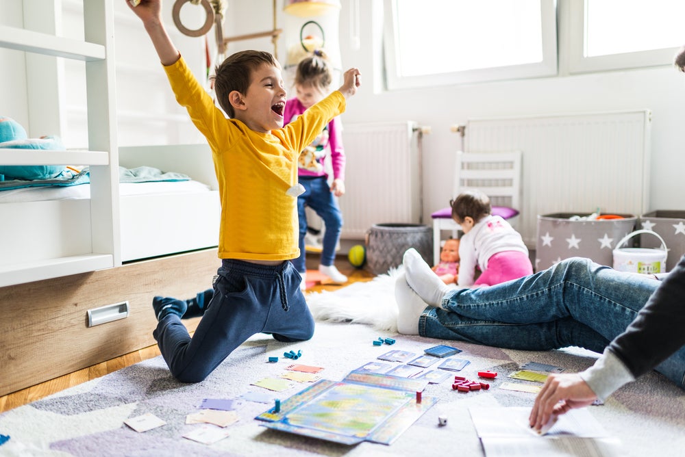 Sala com um adulto, onde só é possível ver suas pernas usando calça jeans e meia. Na mesma sala, estão três crianças. Uma garota com cerca de 1 ano de idade em segundo plano, branca, de cabelos curtos, de costas brincando com uma boneca. Outra garota com cerca de quatro anos, branca, cabelos loiros presos, calça jeans e blusa de manga comprida. Ela parece estar feliz dançando. A criança em destaque é um menino, branco, com cabelos curtos, de braços abertos comemorando uma vitória em um jogo 