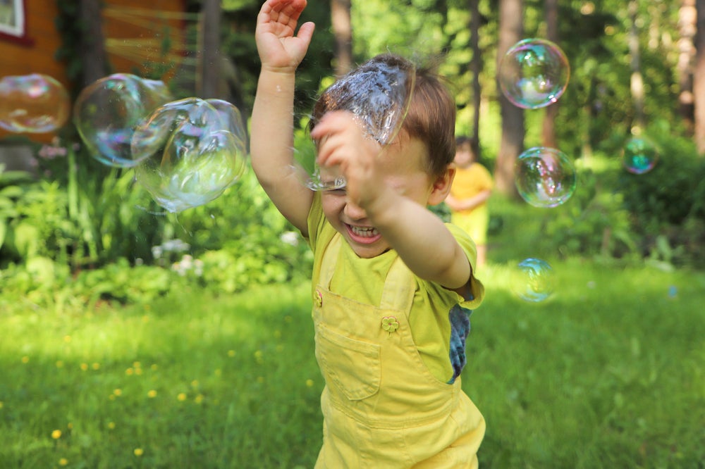 Um menino sorri com as mãos para o alto enquanto passa por bolhas de sabão em um ambiente externo arborizado, com outro menino ao fundo. A foto faz alusão ao tema de brincadeiras ao ar livre. 