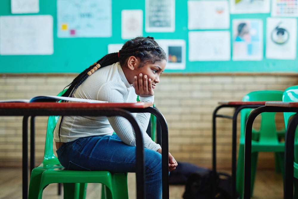 Menina negra, aparentando ter de 8 a 10 anos, de cabelos compridos, presos, está sentada em uma mesa escolar sozinha em uma sala. A mão segura o queixo com expressão chateada. Ela veste calça jeans e blusa verde