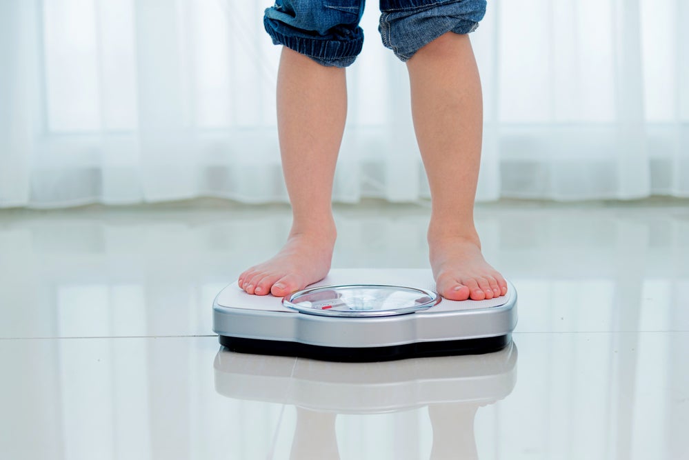 Uma criança com calça jeans dobrada até o joelho se pesa em uma balança digital. Atrás, há uma cortina branca de voil. A foto faz alusão ao tema de transtorno alimentar infantil.