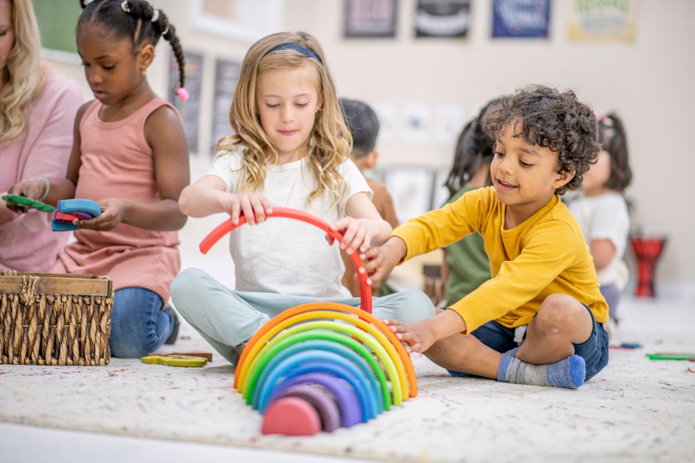  A frente três crianças brincam em uma sala de recreação. Duas delas, uma menina e um menino, estão montando um brinquedo montessori com as cores do arco íris