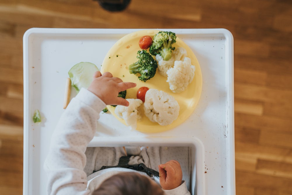 Uma fotografia tirada de cima mostra um bebê em fase de introdução alimentar e seu prato amarelo  com vegetais coloridos a sua frente