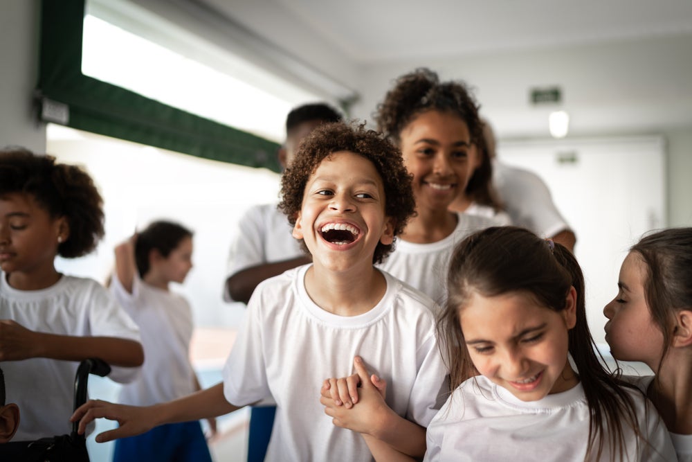 Imagem de um ambiente escolar com várias crianças vestidas de uniforme. O destaque está para um menino gargalhando, bem no centro da foto, de mãos dadas com uma menina