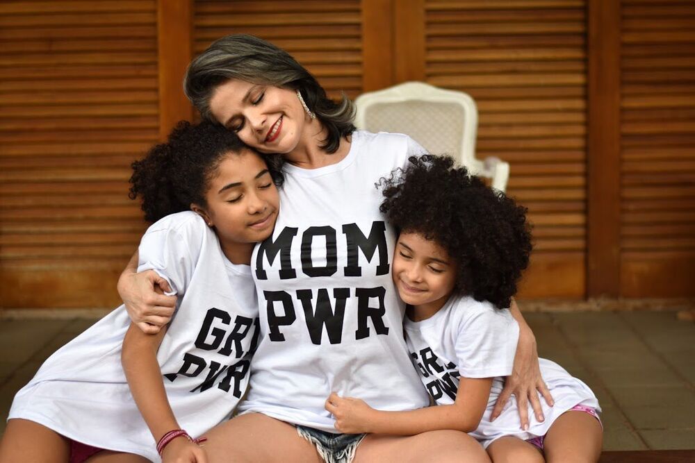 Mãe ao centro da imagem, sentada usando uma camiseta branca com os dizeres "mom pwr", uma filha de cada lado abraçando-a, usando camisetas iguais com os dizerem "grl pwr". Ao fundo uma porta de madeira.
