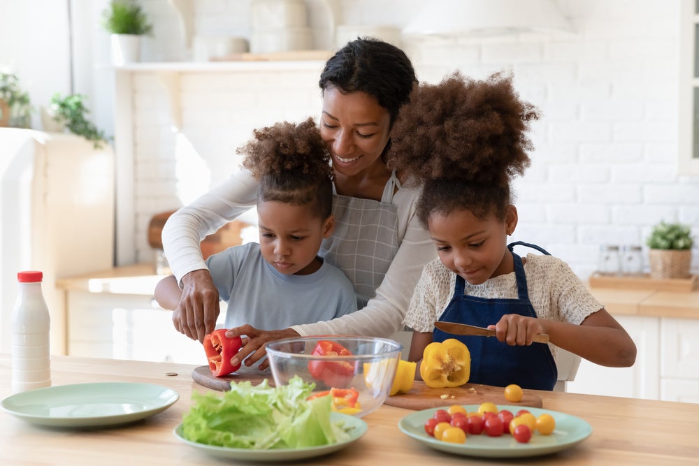  Em uma cozinha, uma mãe ensina os dois filhos a cozinhar. Eles estão em frente a uma bancada de madeira com pratos e ingredientes espalhados, enquanto a mulher ajuda a criança mais nova a cortar um legume.