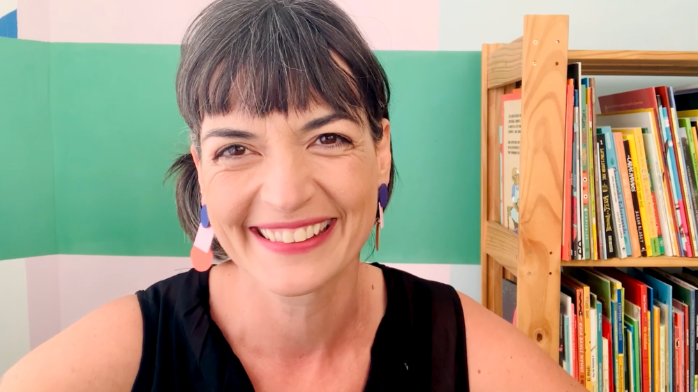 Flávia Scherner sorrindo, ao fundo parede pintada na cor verde e estante com livros coloridos.
