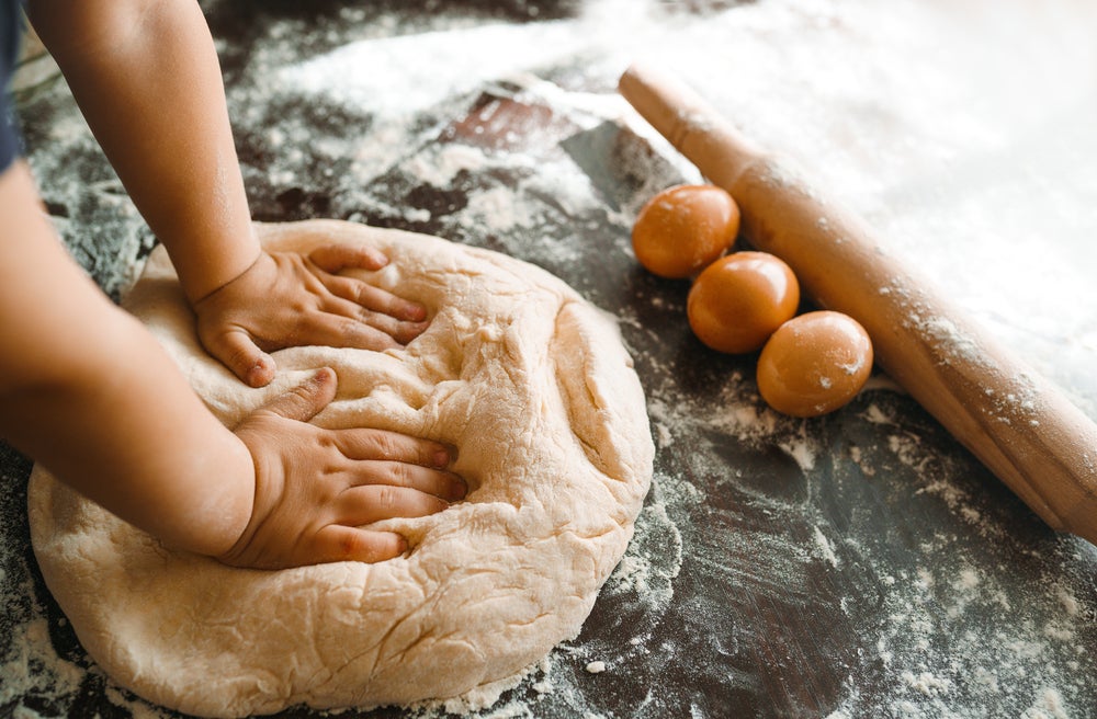 Imagem de mãozinhas de criança amassando um pão caseiro. A frente do pão temos ovos, um rolo de abrir massa e ele está sob uma superfície enfarinhada.