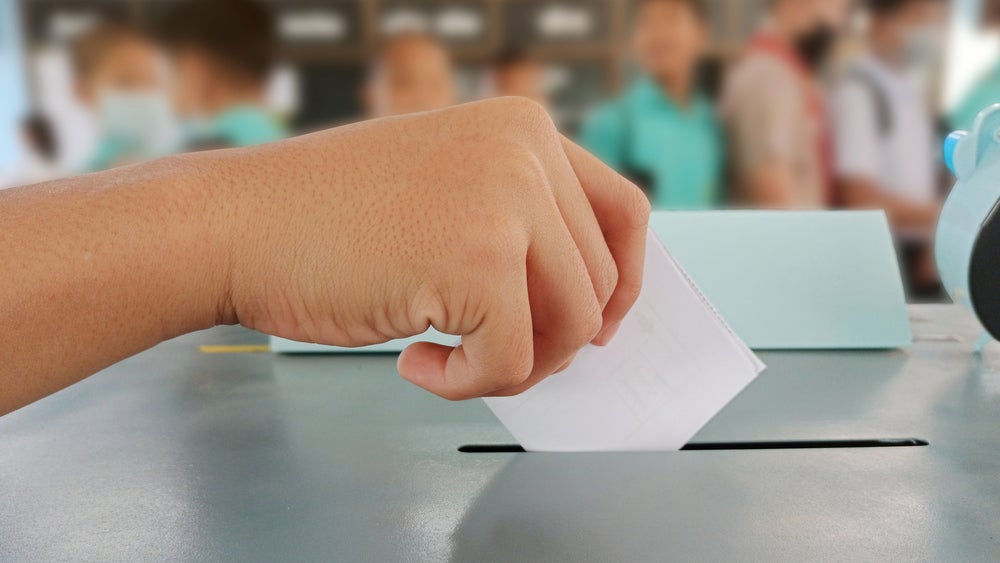 Imagem de uma mão colocando uma cédula de papel dentro de uma urna, para uma votação, uma das coisas que faz parte da política para crianças.
